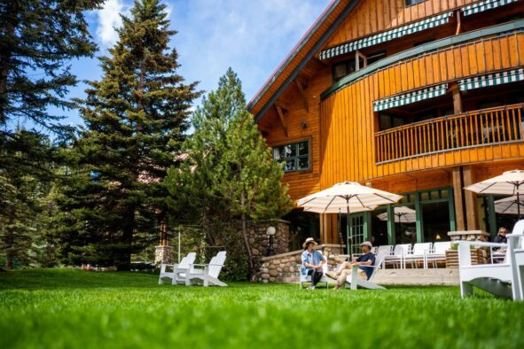 A rustic wooden lodge with guests relaxing on Adirondack chairs on a lush green lawn.